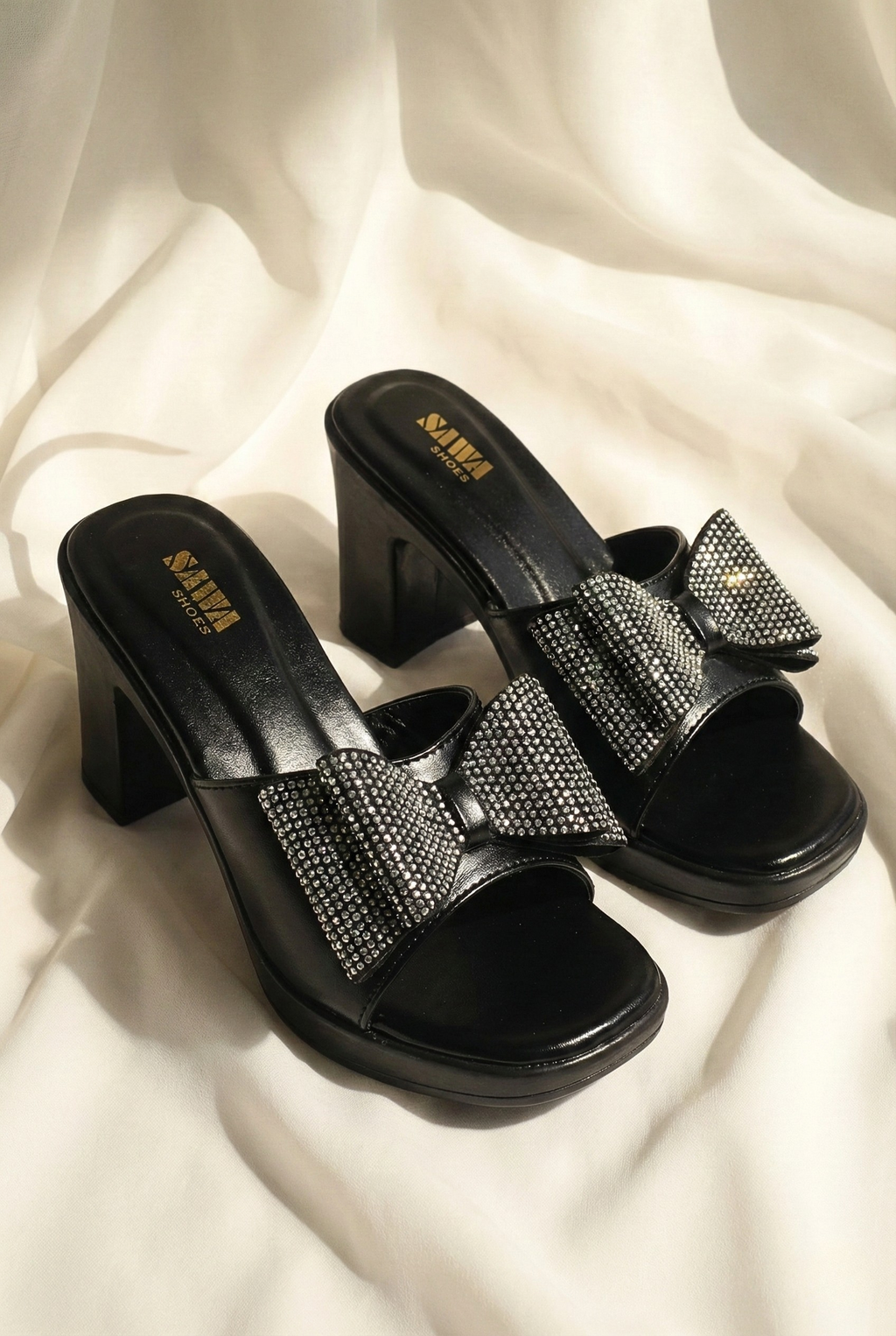 Black high-heeled sandals with rhinestone bows on a white fabric background
