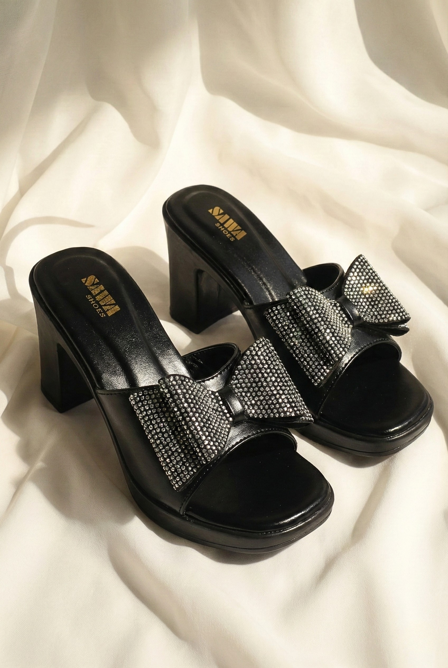 Black high-heeled sandals with rhinestone bows on a white fabric background