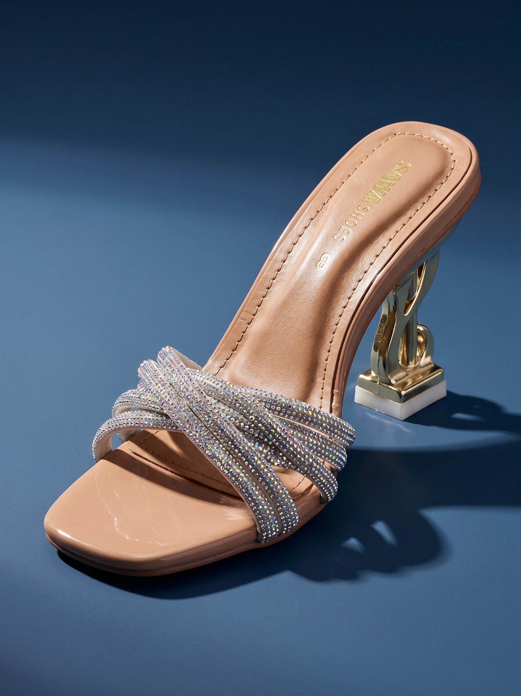 Side view of a nude slip-on sandal featuring a 3-inch gold sculptural heel and shimmering crystal-encrusted straps against a light blue background.