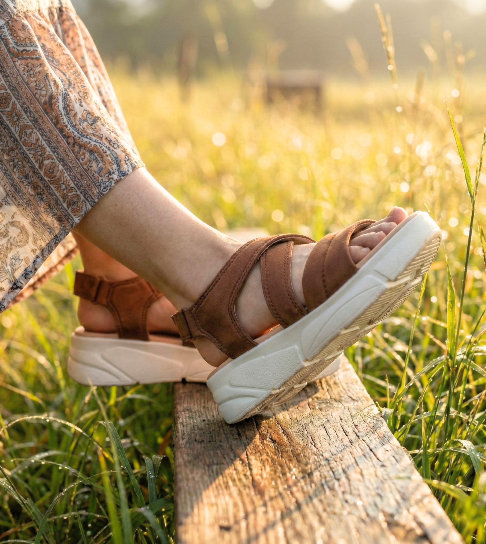 Side profile of tan strappy platform sandals with a cushioned white wedge sole, worn by a woman sitting on a wooden bench in a sunlit meadow
