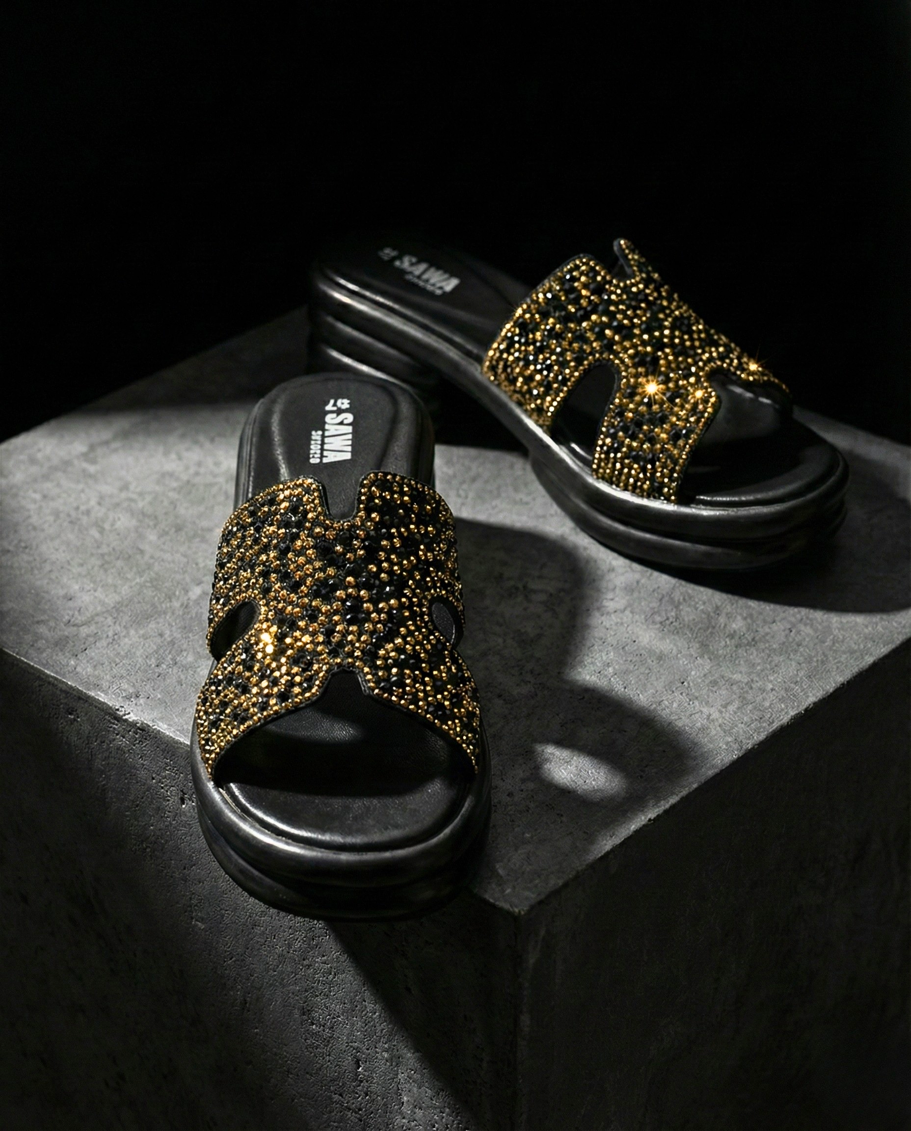Close-up of black SAWA SHOES platform slide sandals featuring a wide strap encrusted with gold and black rhinestones on a textured gray stone surface