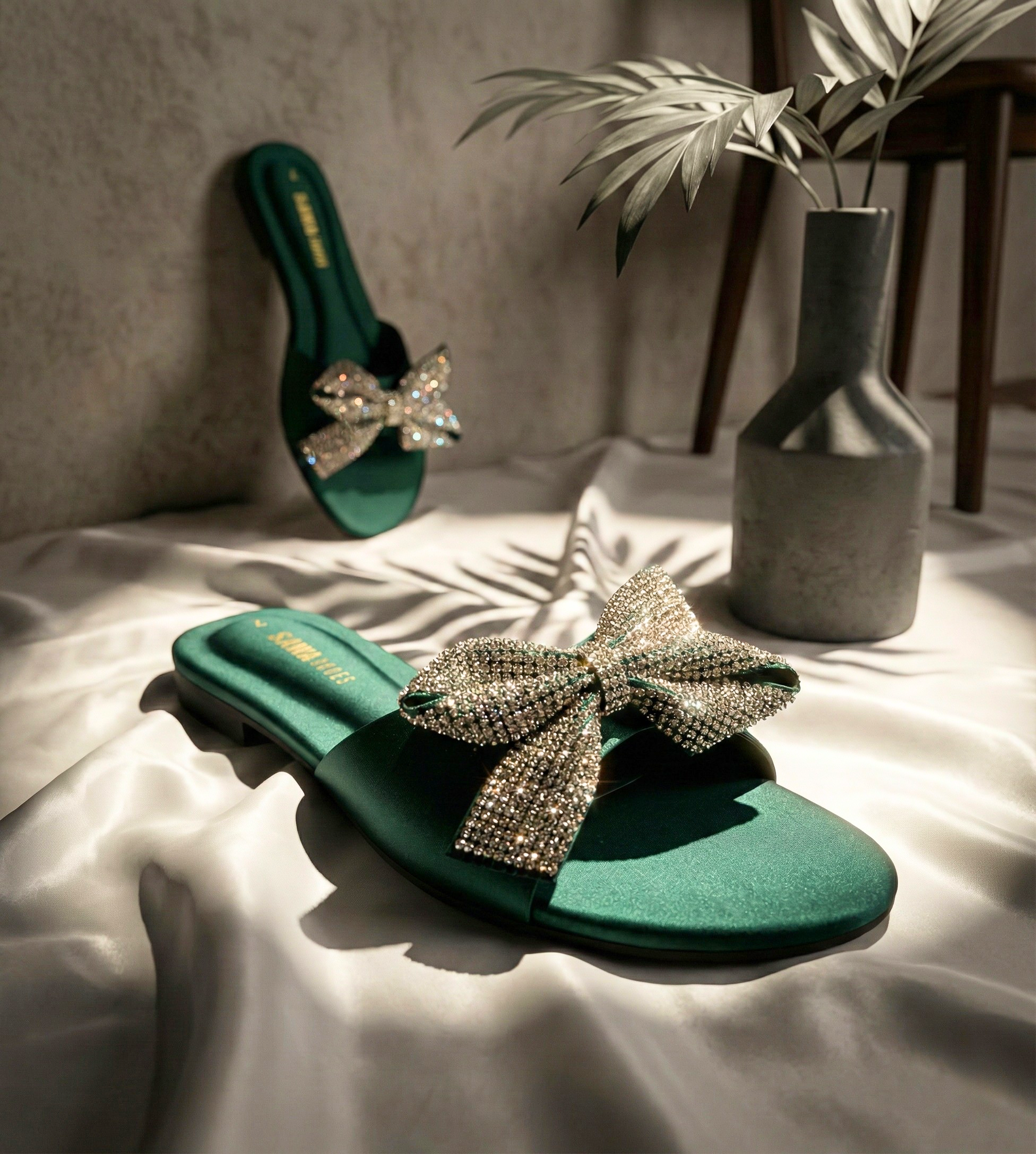 Emerald green satin slide with a large crystal-encrusted bow on a white draped fabric background.