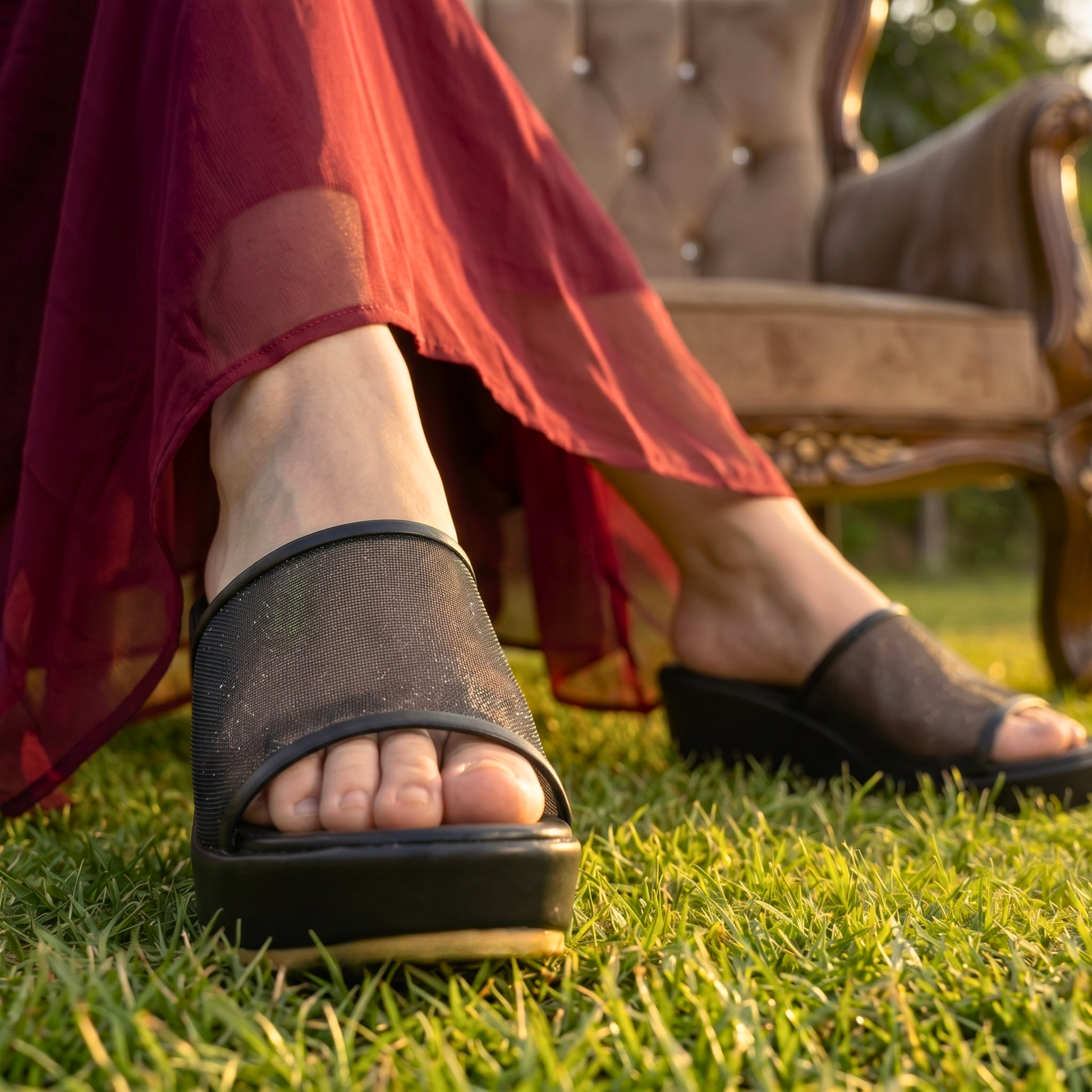 a close-up shot of a person's feet wearing black mesh strap wedge sandals while walking on green grass during sunset