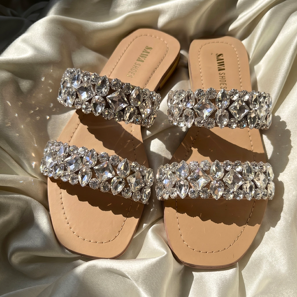 Pair of nude-colored flat slide sandals with two wide straps heavily embellished with shimmering, multi-shaped clear crystals, displayed on a draped silk fabric and worn on feet with a champagne-colored dress.