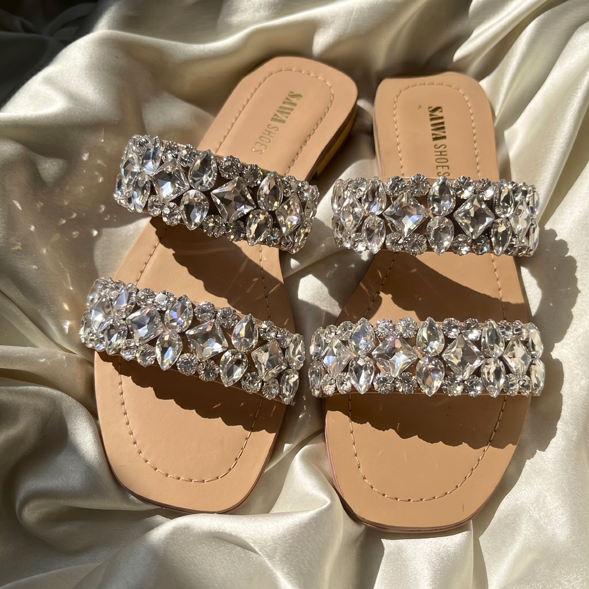 Pair of nude-colored flat slide sandals with two wide straps heavily embellished with shimmering, multi-shaped clear crystals, displayed on a draped silk fabric and worn on feet with a champagne-colored dress.