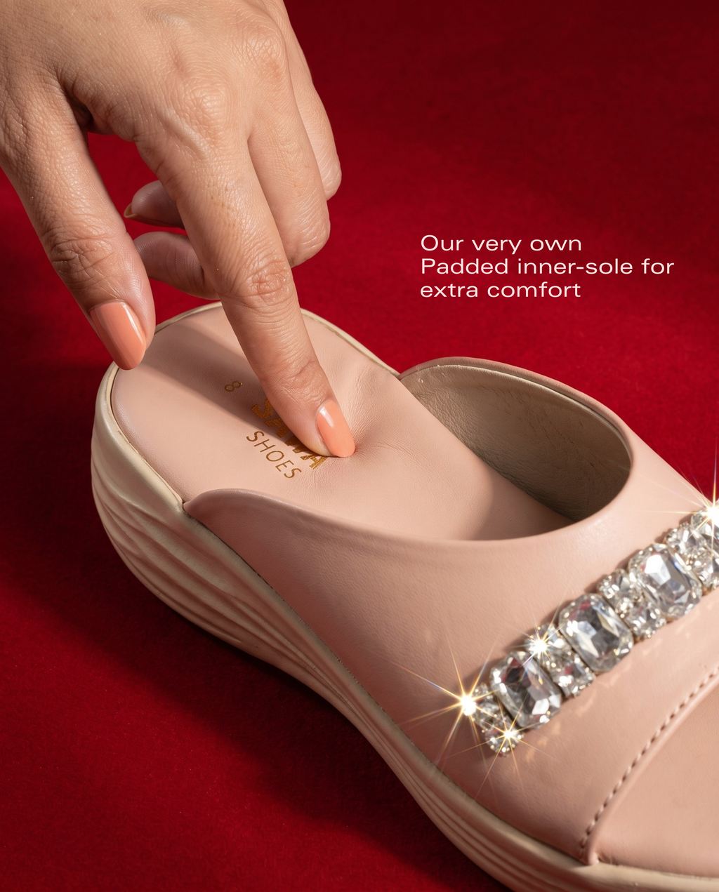 A close-up of a nude pink wedge slip-on shoe with a jeweled embellishment on the side, shown on a red background. A hand is pressing into the padded insole, with text that reads 'Our very own Padded inner-sole for extra comfort'.