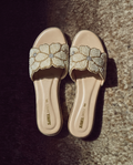 Pair of cream flat slides with floral pearl embellishments and gold accents angled on a textured backdrop.