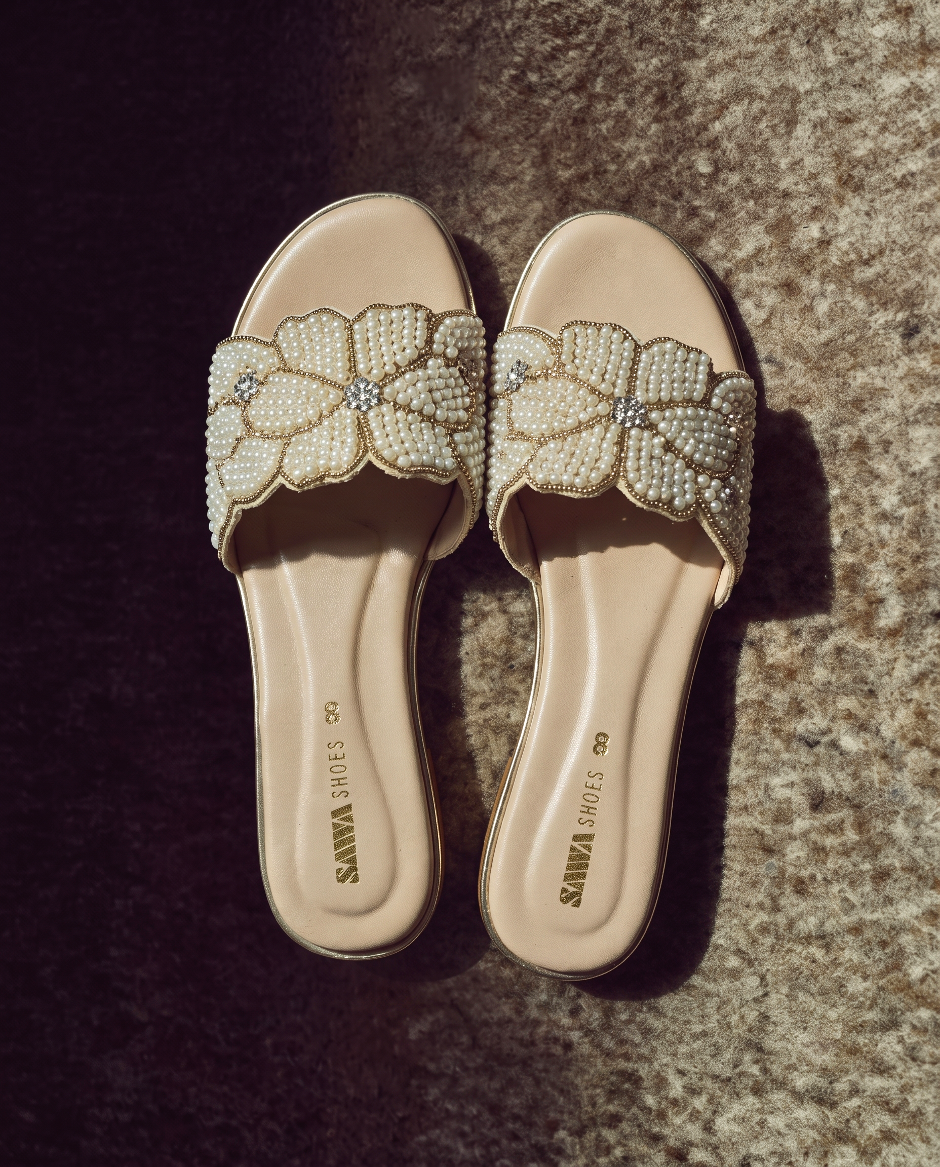 Pair of cream flat slides with floral pearl embellishments and gold accents angled on a textured backdrop.