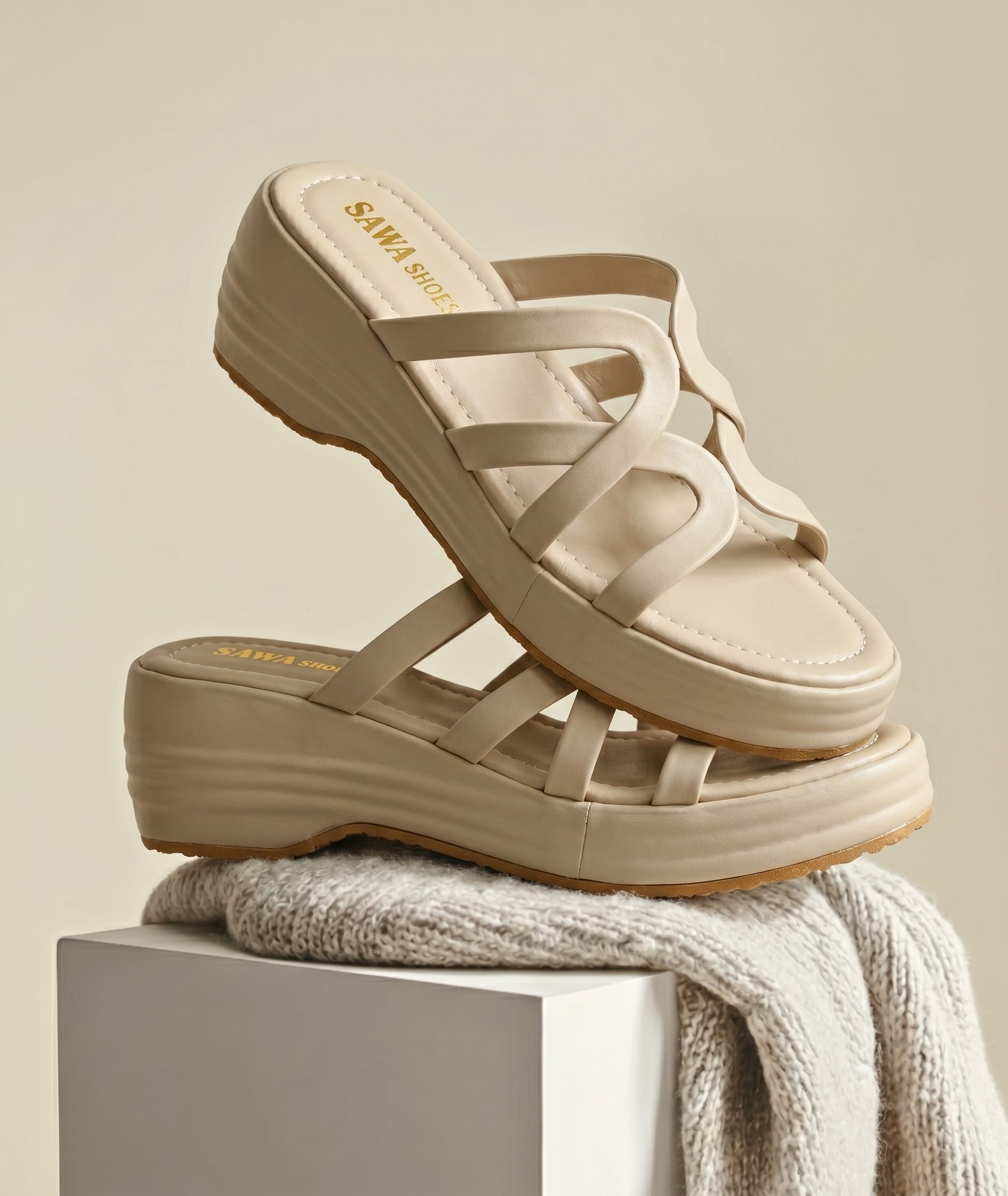 A pair of neutral-toned platform wedge sandals stacked on a white pedestal, highlighting the interlocking upper straps.