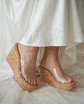 Woman wearing clear strap cork platform wedge sandals with a 3.8-inch heel and tan cushioned footbed paired with a white satin dress