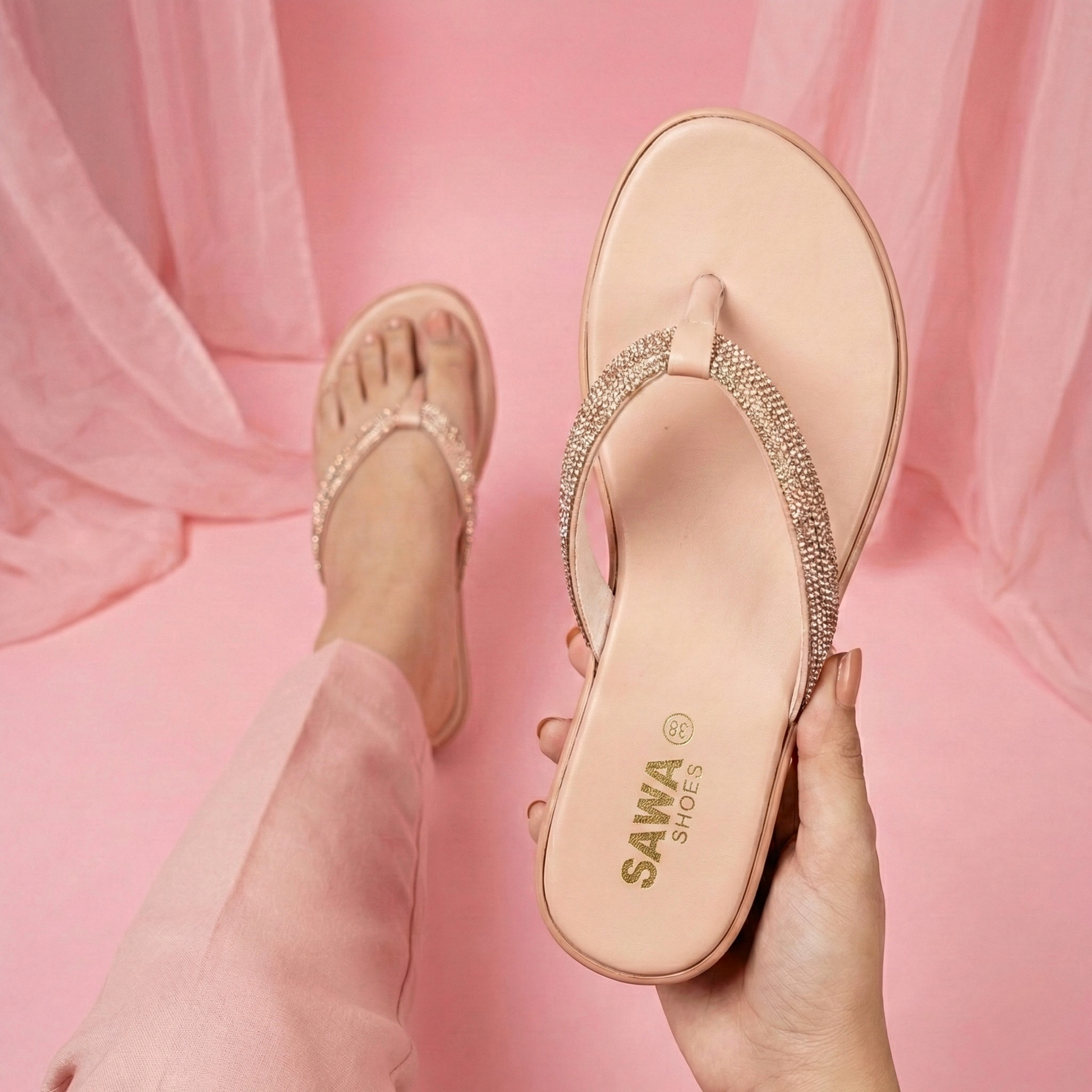 Pink sandal with rhinestone details held by a hand against a pink fabric background