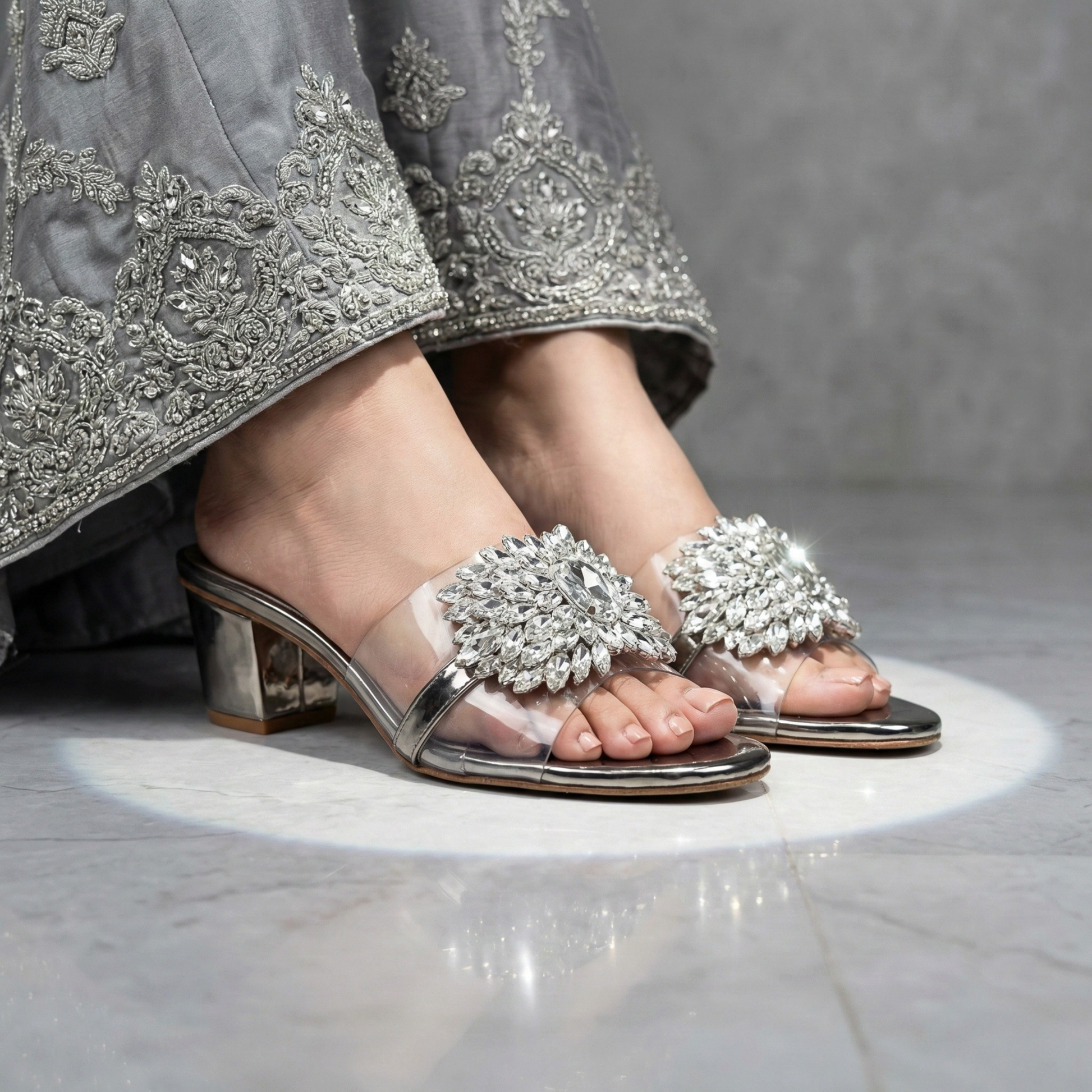 Side view of a person wearing silver block-heel mules with a clear PVC strap and large crystal floral embellishment paired with an embroidered silver dress