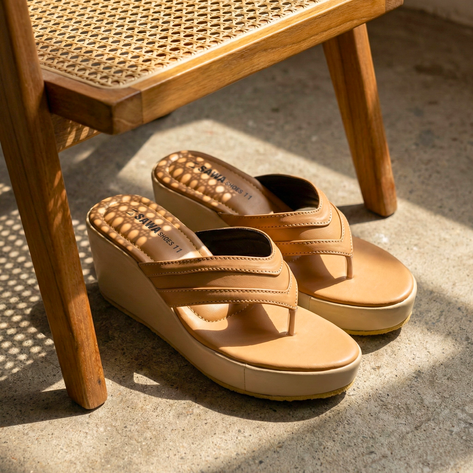 Minimalist tan platform wedge sandals with a unique triple-stitched V-shaped thong strap, shown in natural sunlight on a stone floor.