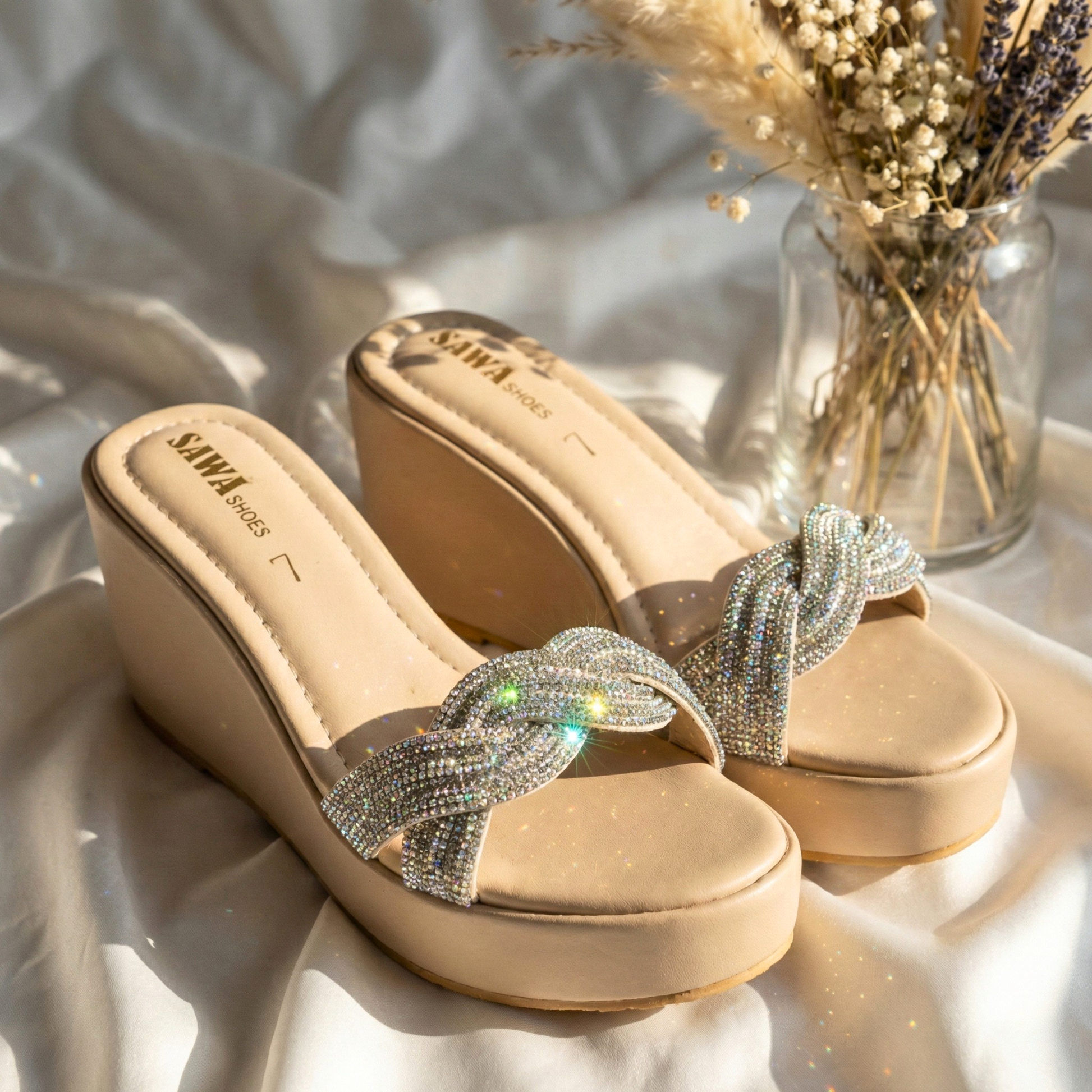 Sparkling nude platform wedges with crystal-encrusted braided straps, displayed on a white silk backdrop with dried flowers