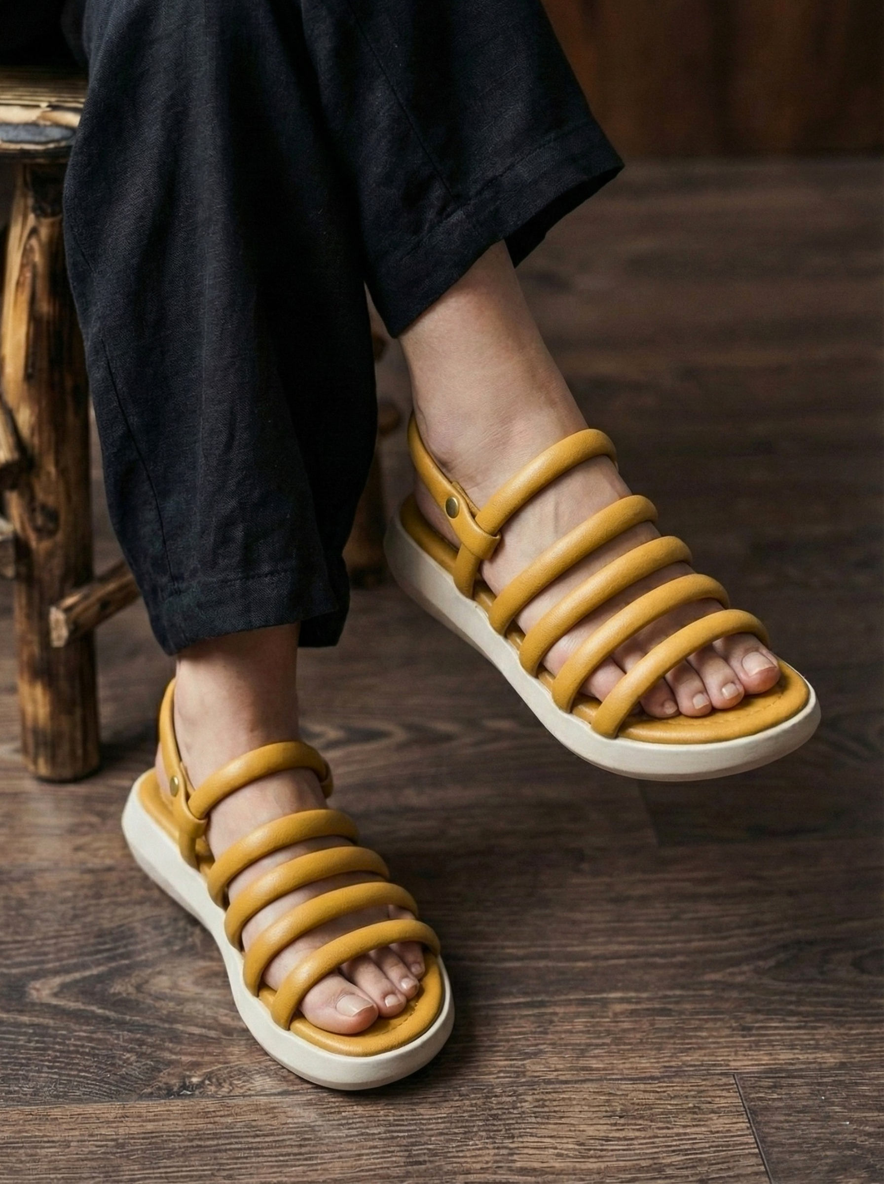 Woman wearing mustard yellow ochre sandals with puffy tubular straps and white flatform sole, sitting on a wooden stool. Stylish comfortable ladies footwear for summer in Pakistan