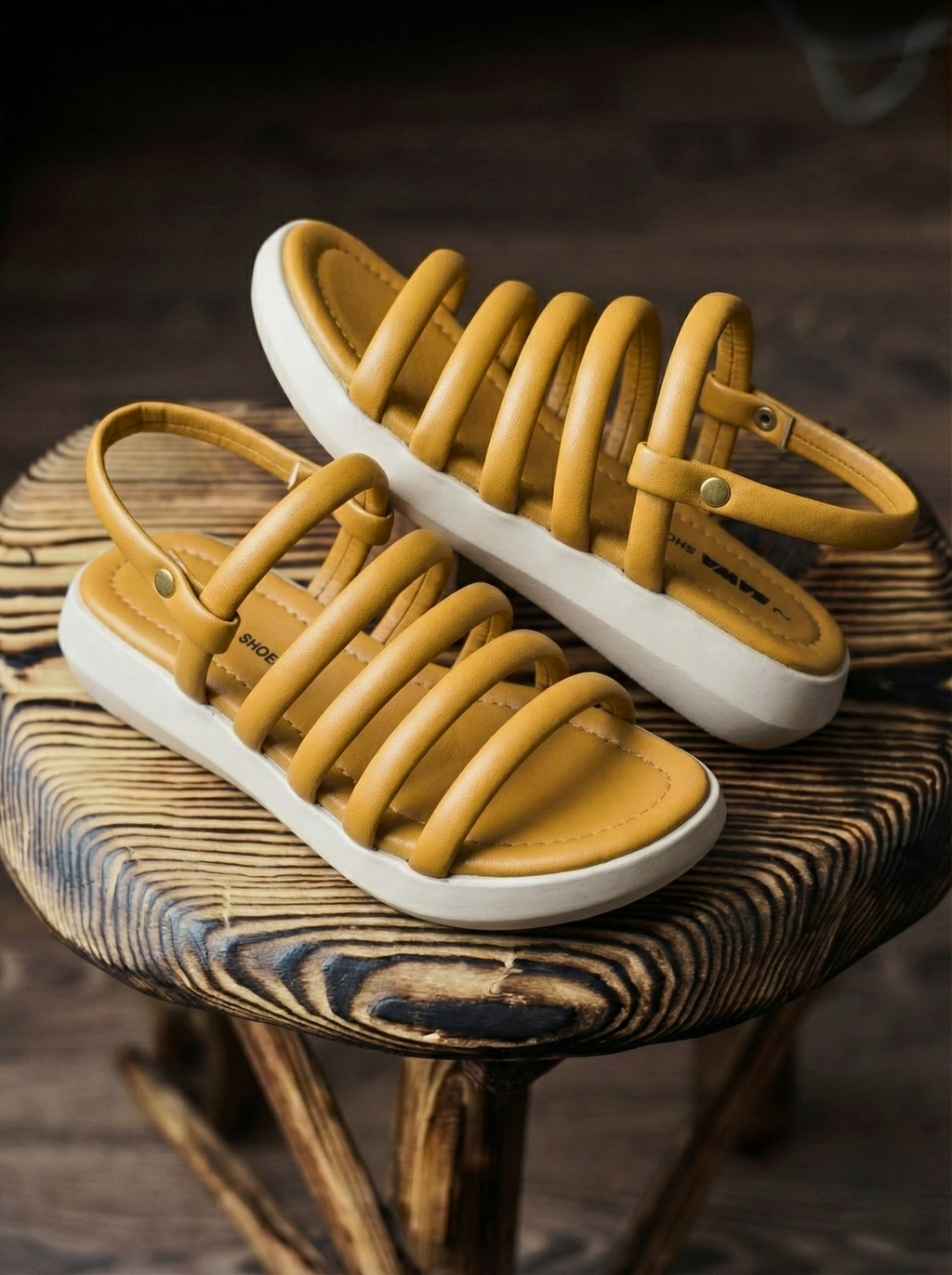 Woman wearing mustard yellow ochre sandals with puffy tubular straps and white flatform sole, sitting on a wooden stool. Stylish comfortable ladies footwear for summer in Pakistan