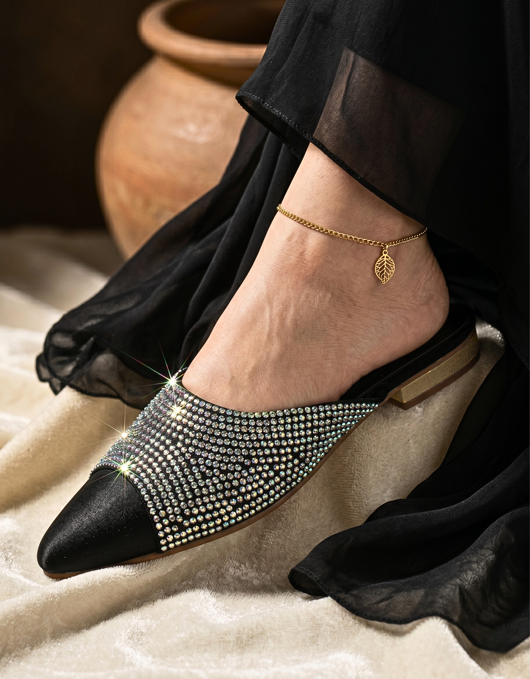 Side view of a person wearing the shimmering rhinestone black mule shoe, paired with an elegant gold leaf anklet charm and a flowing black fabric dress