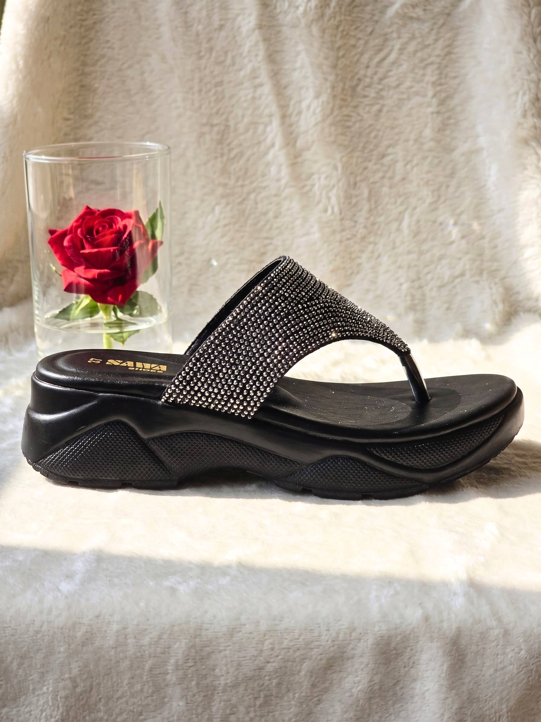Black platform wedge sandals with shimmering rhinestone straps placed next to a red rose in a glass