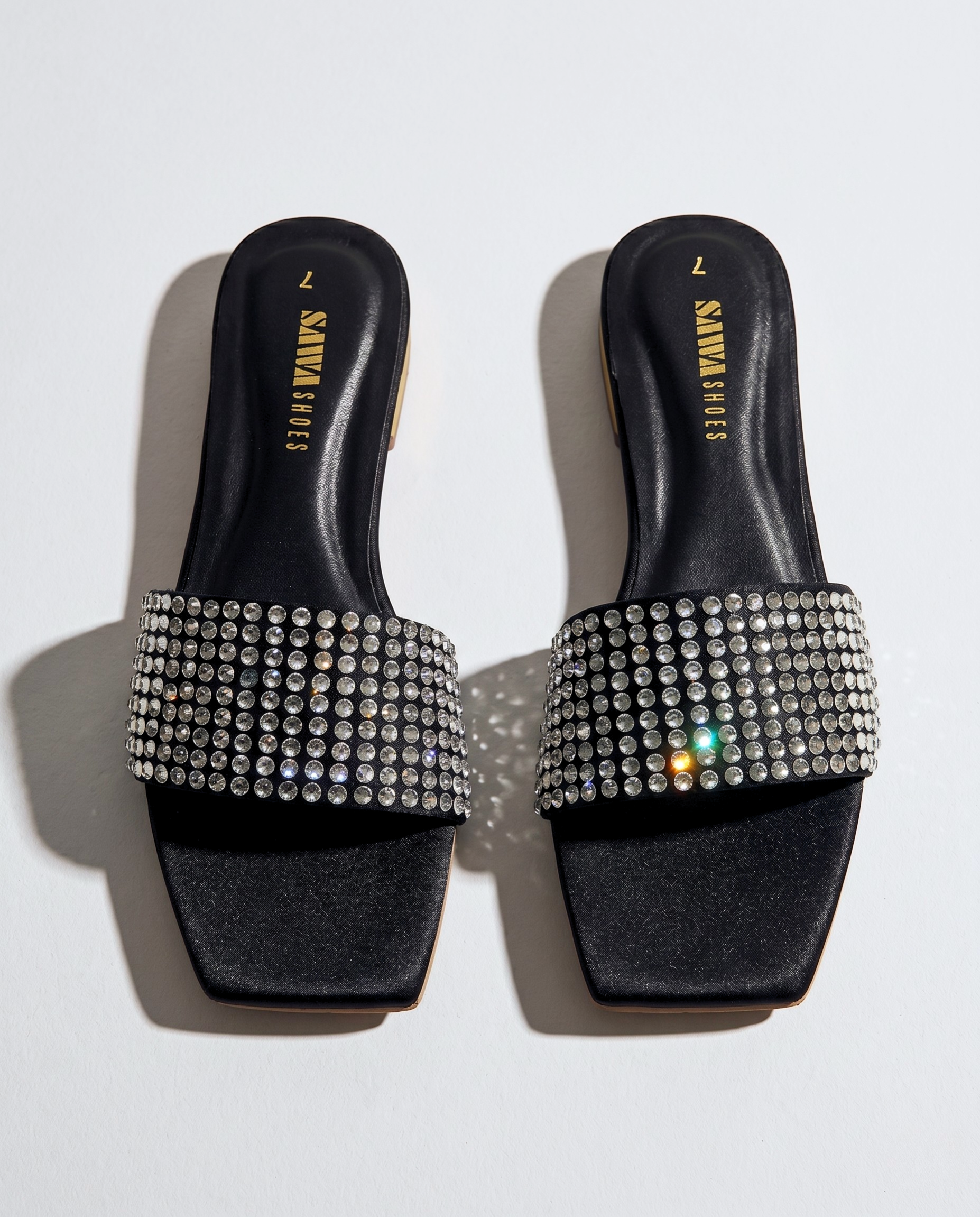 Top-down view of a pair of black square-toe slide sandals with wide straps covered in glittering silver crystals on a white background