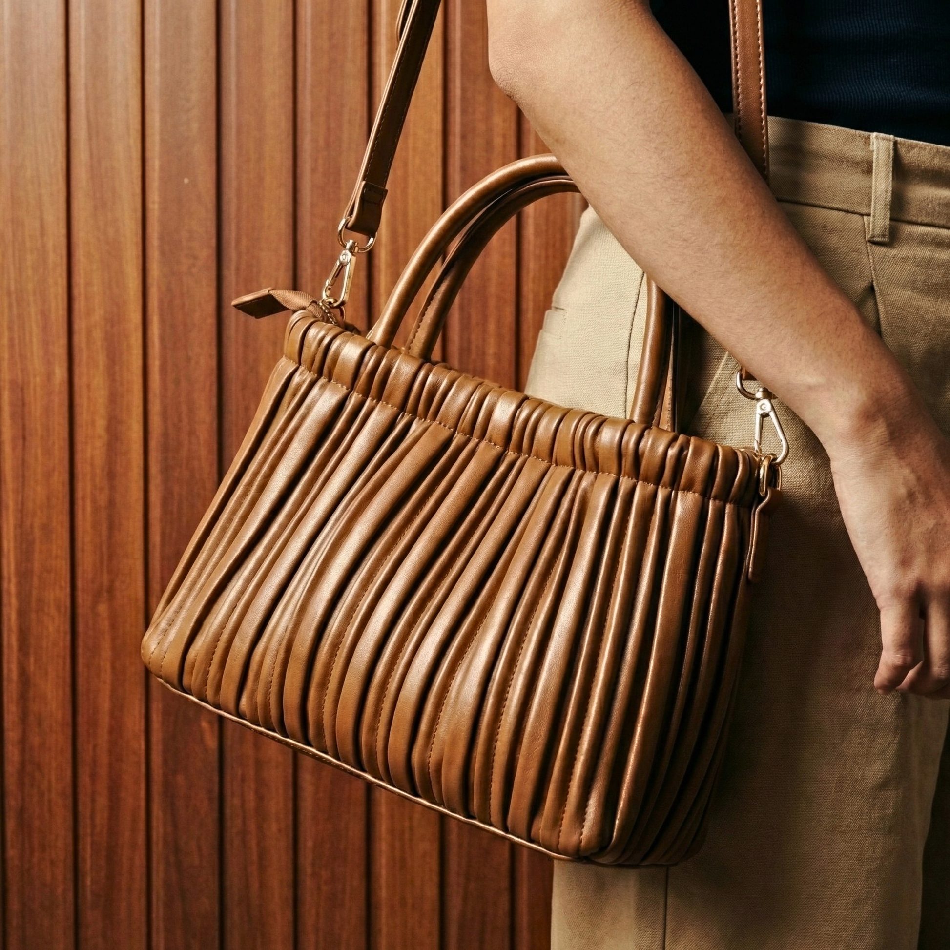 Brown pleated handbag held by a person against a wooden panel background