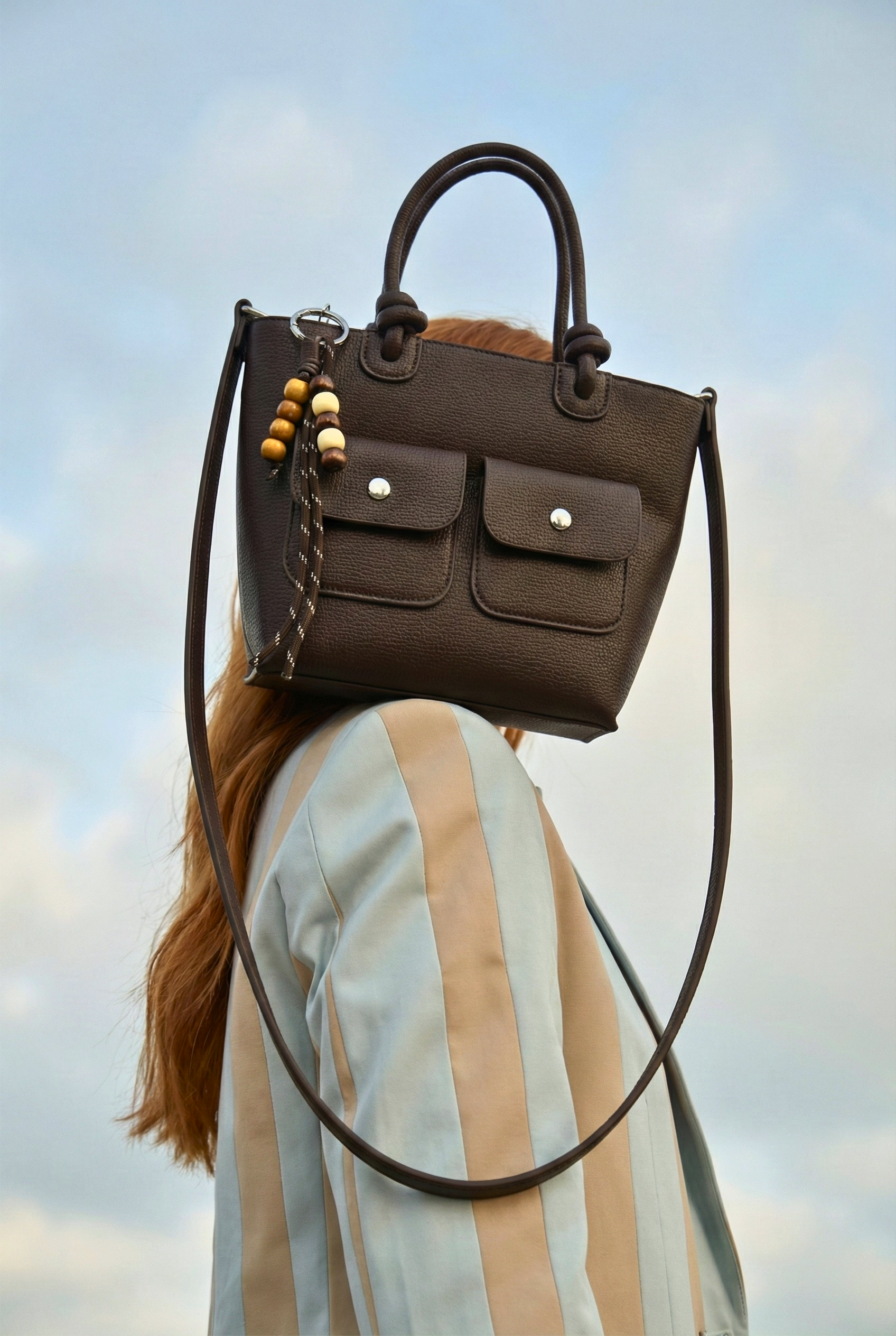 Front view of a dark chocolate brown pebbled leather handbag featuring two flap pockets with silver snaps and a decorative wooden bead charm