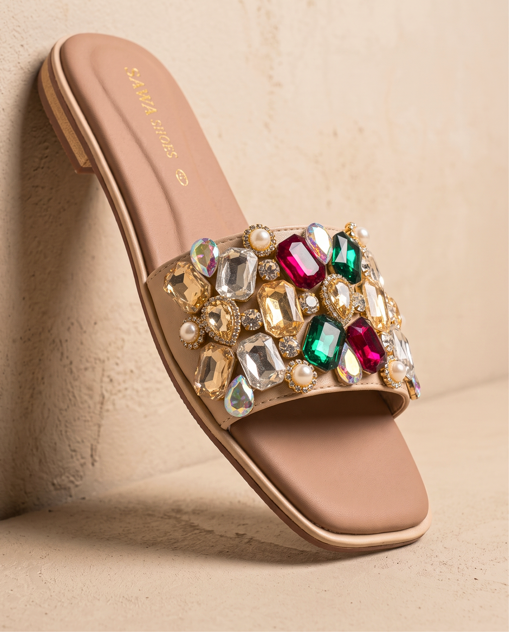 A pair of nude flat slide sandals by SAWA SHOES with a modern square toe. The wide top strap is heavily embellished with large, multicolored faceted gems in shades of emerald, ruby, clear, and gold, accented by small white pearls.