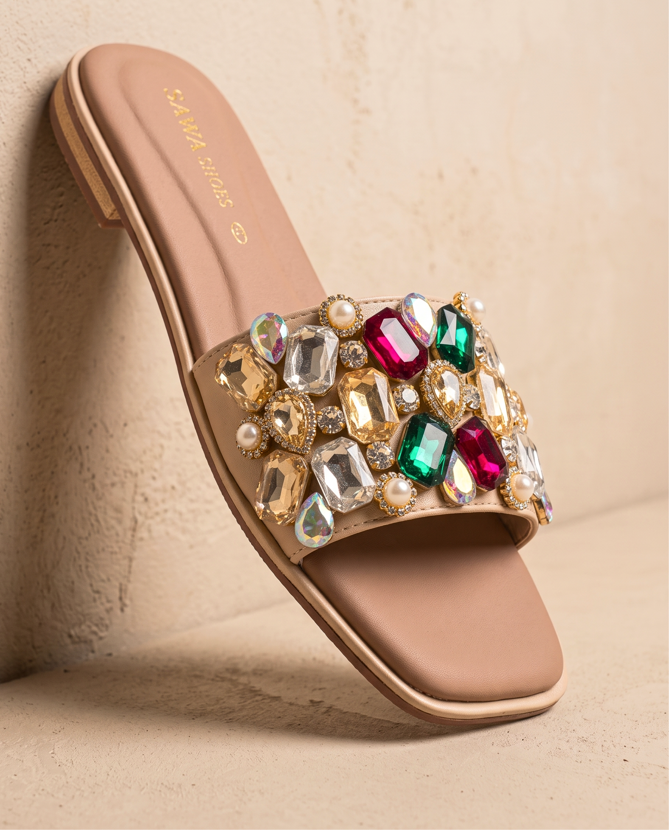 A pair of nude flat slide sandals by SAWA SHOES with a modern square toe. The wide top strap is heavily embellished with large, multicolored faceted gems in shades of emerald, ruby, clear, and gold, accented by small white pearls.