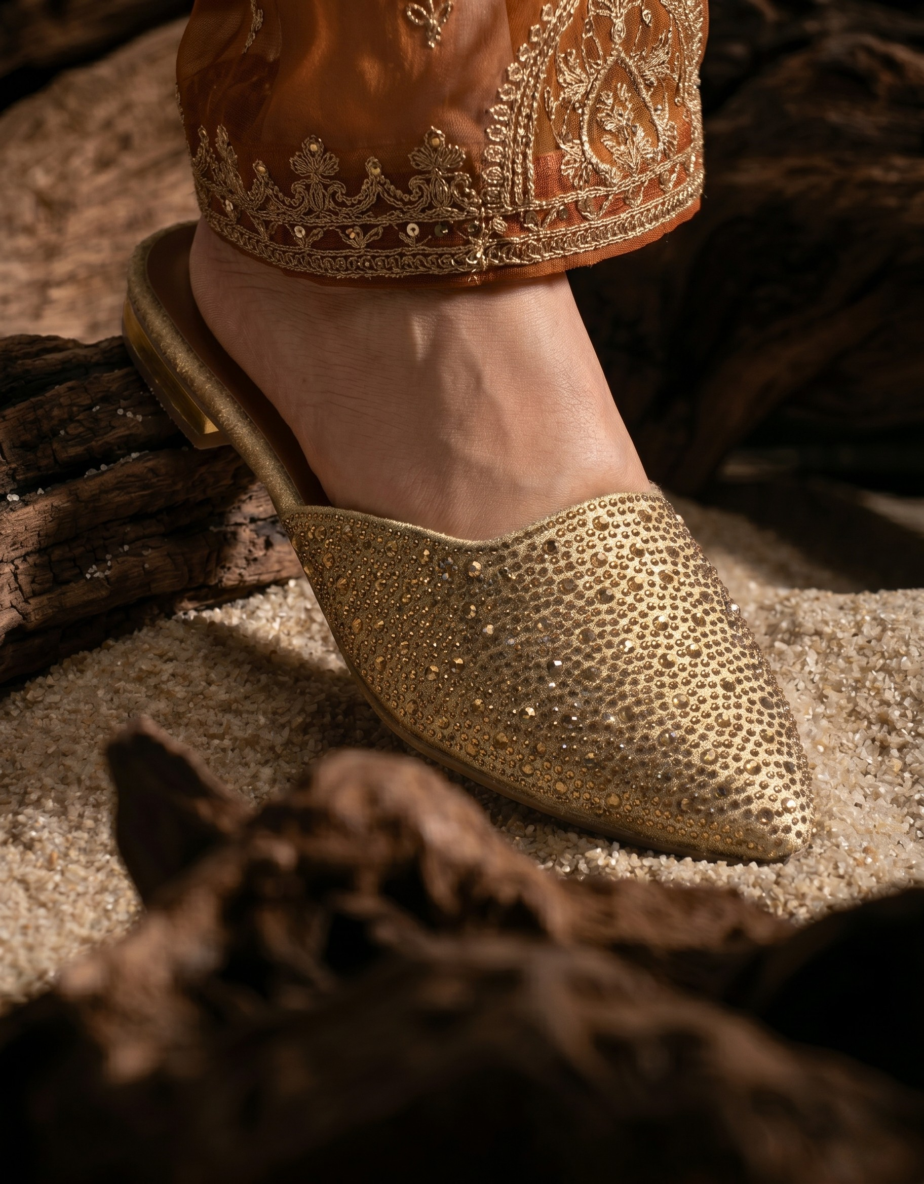 Close-up of a person wearing gold rhinestone-embellished pointed-toe mules paired with an embroidered bronze-orange traditional outfit.