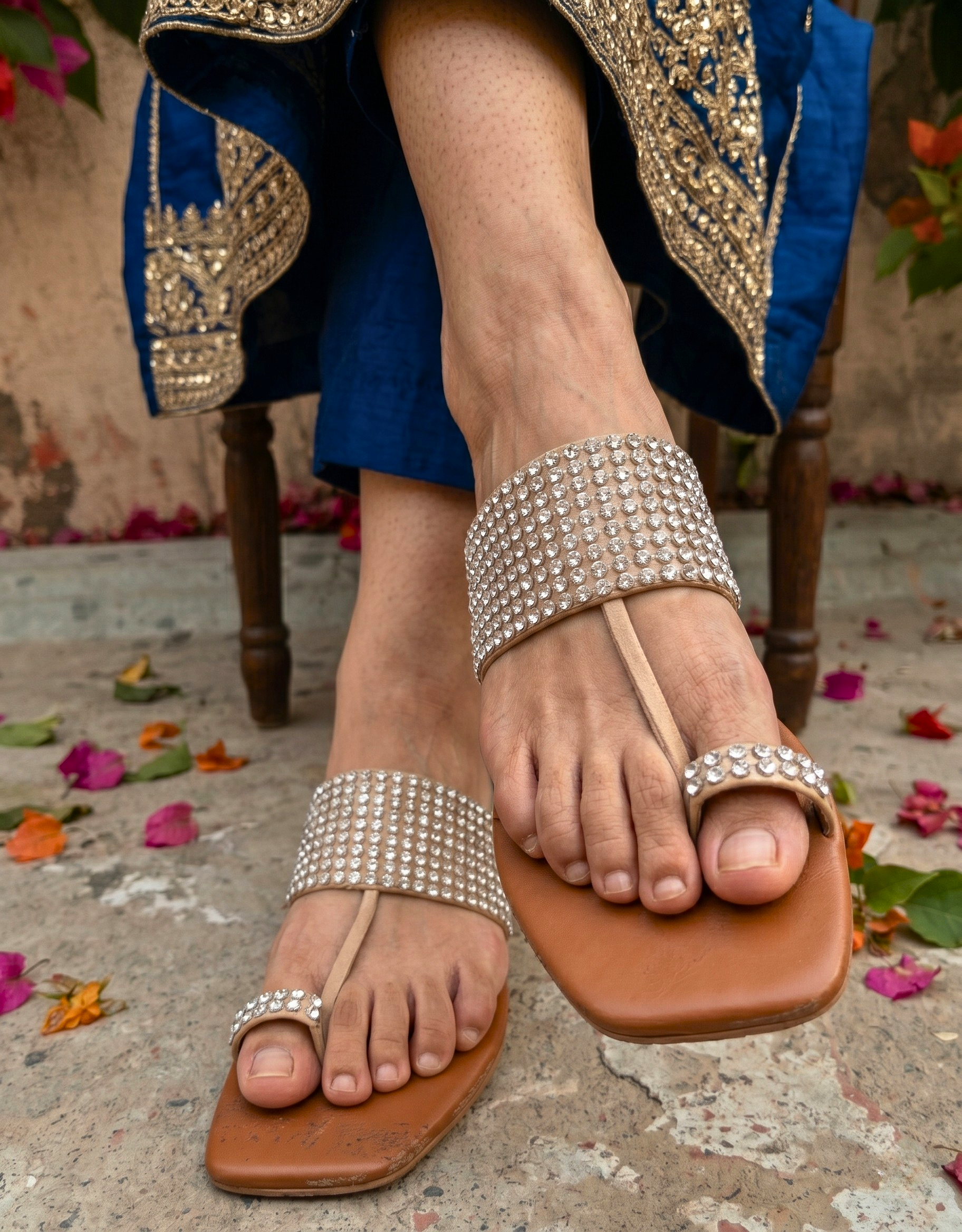 A pair of tan Kolhapuri-style flat sandals by Sawa Shoes featuring wide upper straps and toe loops heavily embellished with sparkling rows of rhinestones, set against a rustic concrete background with pink bougainvillea flowers.