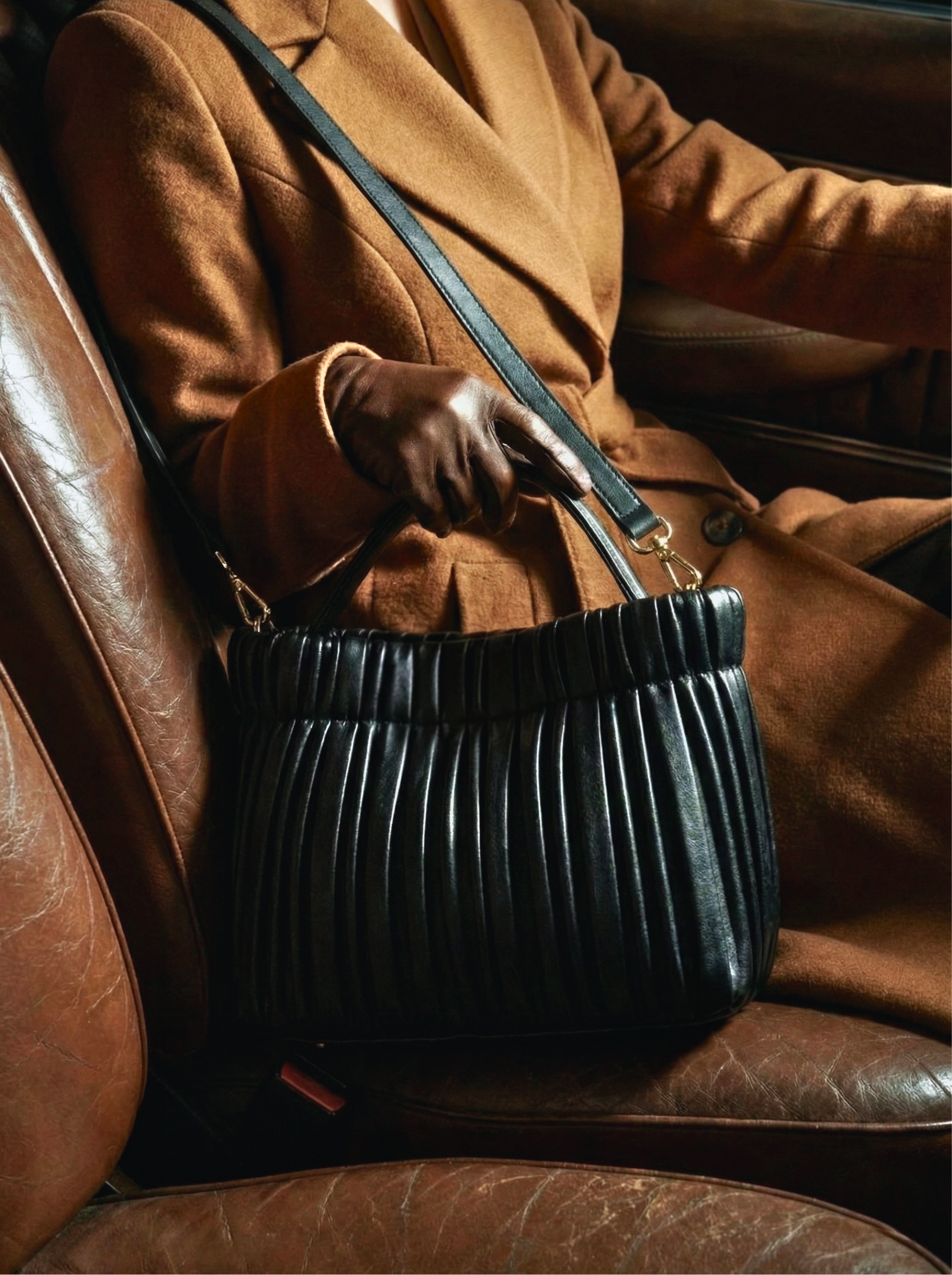 A black pleated leather handbag held by a hand wearing a brown leather glove against a tan wool coat