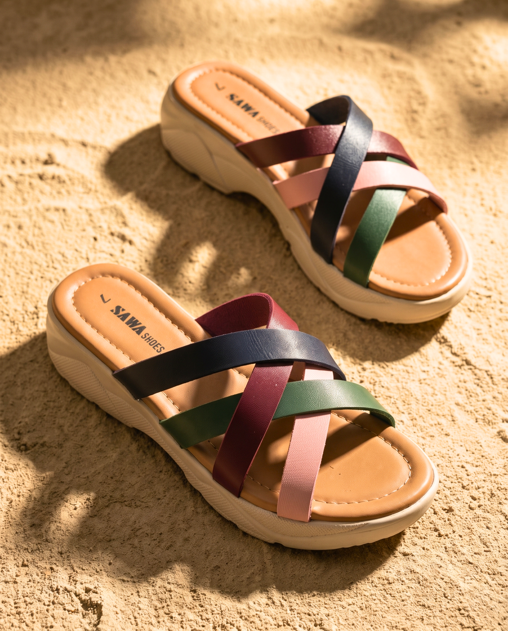 A pair of SAWA SHOES multi-strap platform sandals on sand. One shoe is leaning against a warm, sunlit wall, while the other rests flat in the foreground.