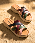 A pair of SAWA SHOES multi-strap platform sandals on sand. One shoe is leaning against a warm, sunlit wall, while the other rests flat in the foreground.