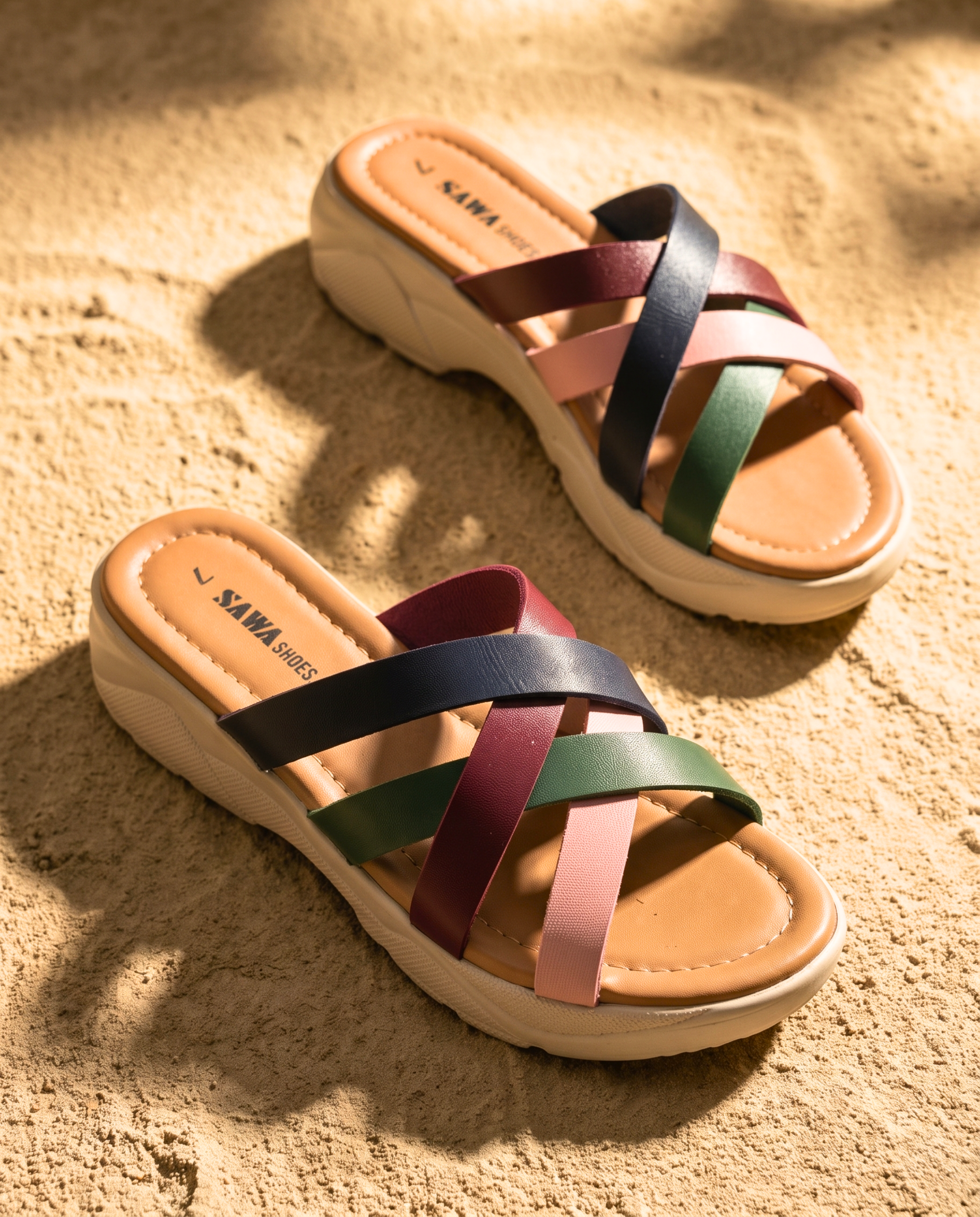 A pair of SAWA SHOES multi-strap platform sandals on sand. One shoe is leaning against a warm, sunlit wall, while the other rests flat in the foreground.