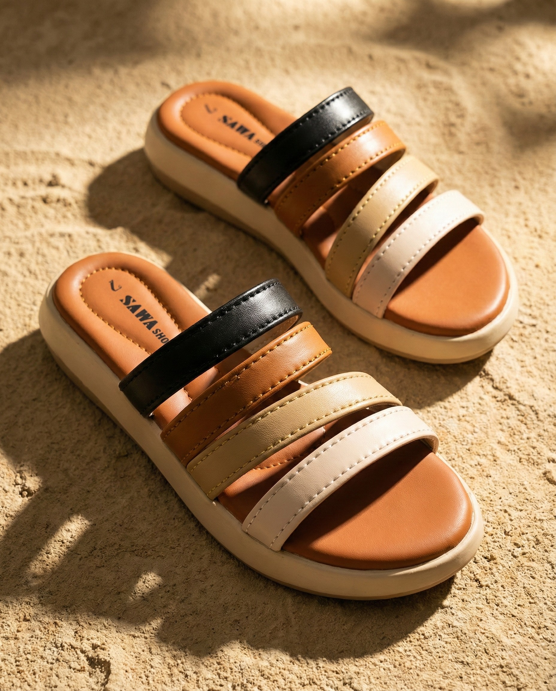 A pair of SAWA brand medicated slides on a sandy background. The sandals feature four horizontal straps in black, tan, beige, and cream colors with a thick, cushioned cream-colored platform sole