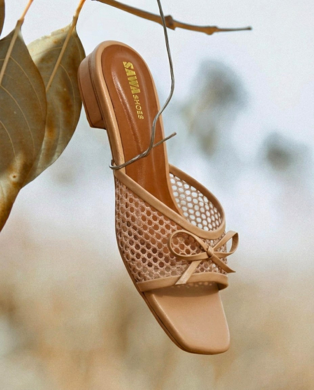 Top view of SAWA Shoes nude mesh bow mule featuring a square toe and gold logo on the footbed, laid on concrete