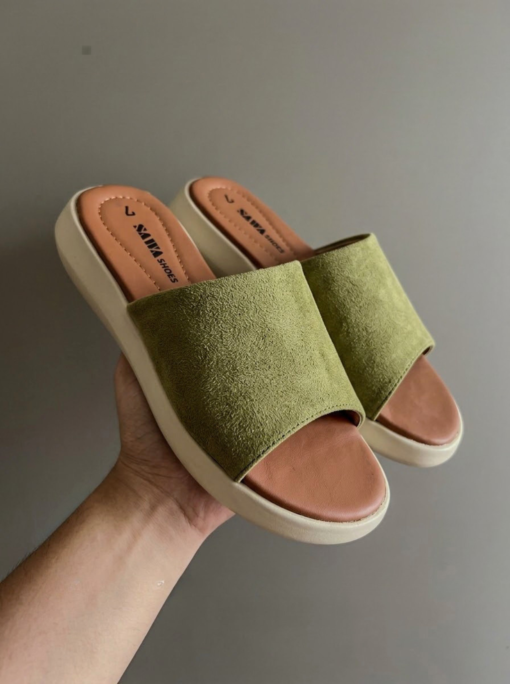 A pair of green slide sandals with tan footbeds and off-white soles, held in a hand against a plain gray background.