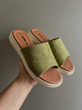 A pair of green slide sandals with tan footbeds and off-white soles, held in a hand against a plain gray background.