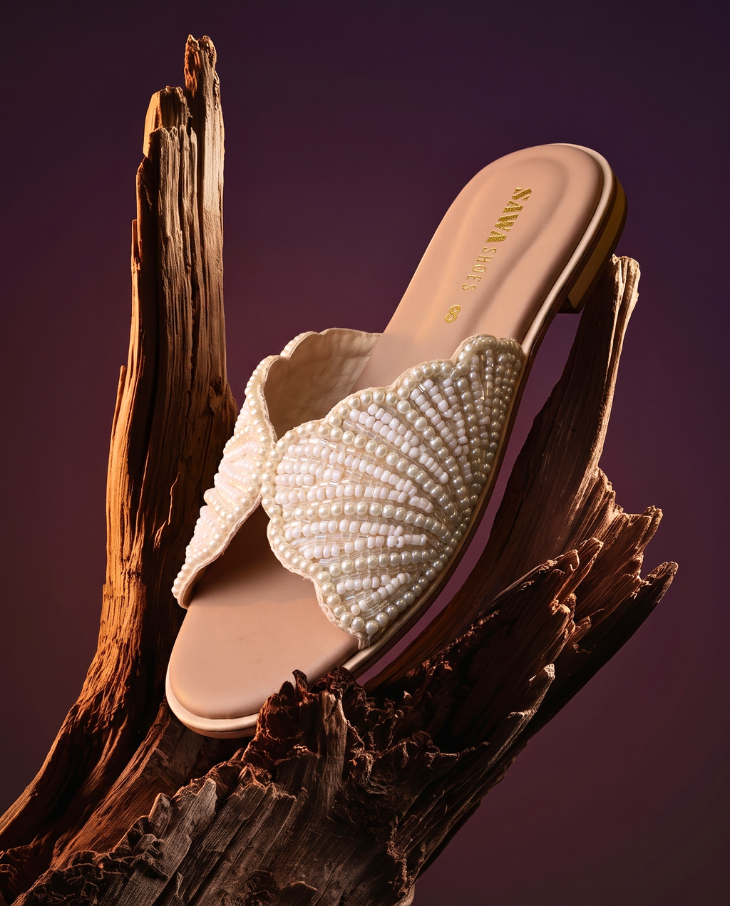 A pair of nude flat slide sandals by Sawa Shoes resting on a piece of decorative driftwood. The wide top strap features a scalloped edge and is entirely covered in small white pearls arranged in a textured shell pattern.