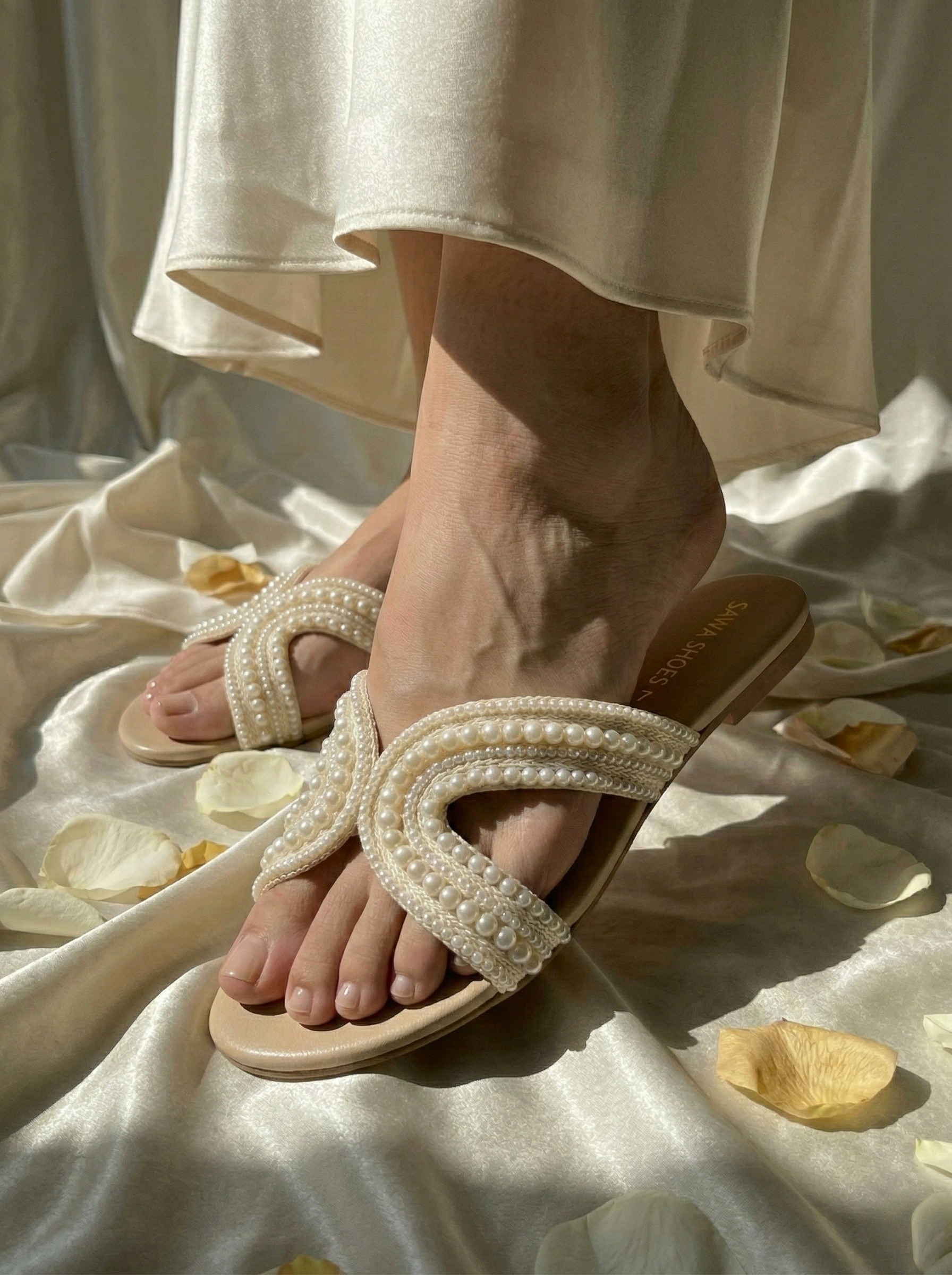 Close-up of a person wearing Sawa Shoes Whispers of Pearl ivory flat sandals with crossover pearl-embellished straps on a cream satin background with rose petals.