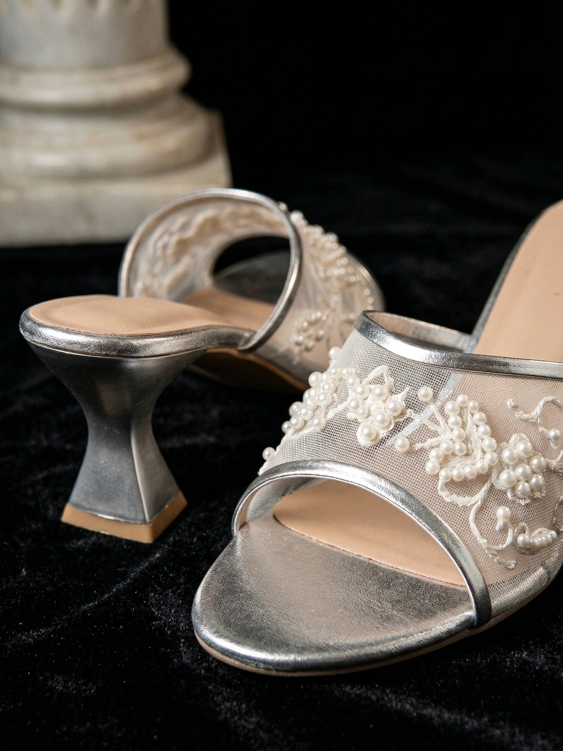 Side view of metallic silver mesh mules showing the architectural flared block heel and pearl embellishments