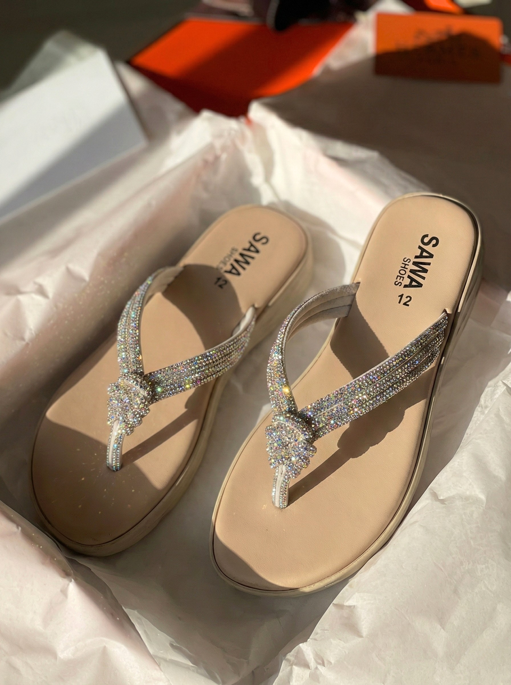 A pair of beige Sawa brand wedge thong sandals featuring straps covered in iridescent crystals with a decorative knot detail, resting in a white box