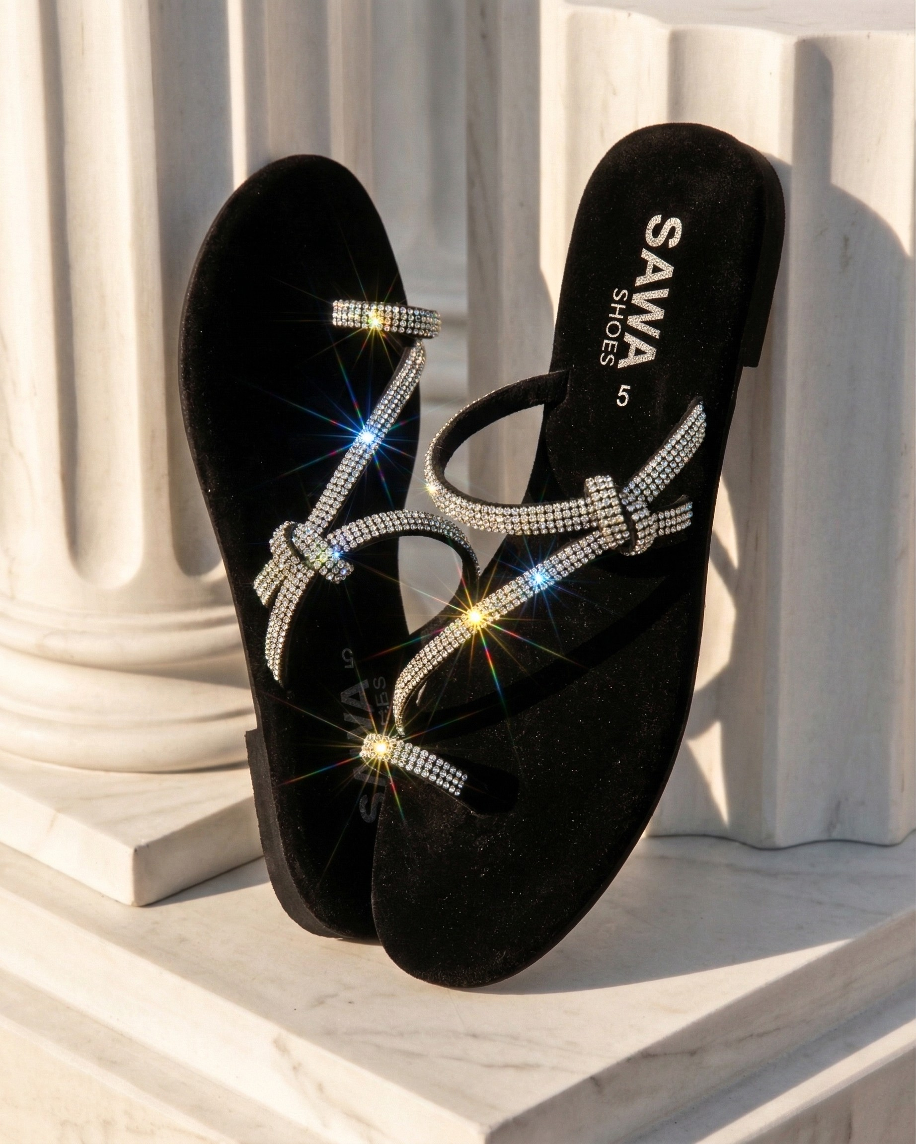 SAWA black velvet slides with sparkling rhinestone straps and a decorative knot detail, displayed against white marble columns
