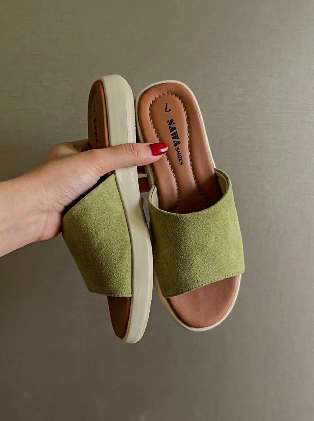 A pair of olive green slide sandals with cream-colored soles, held by a hand with red nail polish against a neutral gray background. The brand name 'SALVADOR' is visible on the insole.